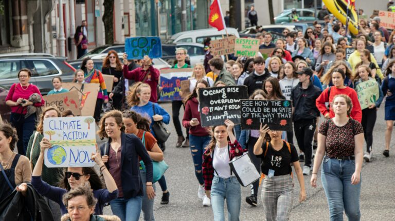 A large group of people protesting against global warming and in defense of the environment. They are marching in a city, and some carry signs with messages saying: “Climate action now,” “Oceans rise & overflow,” and “It ain't right & it ain't natural.”