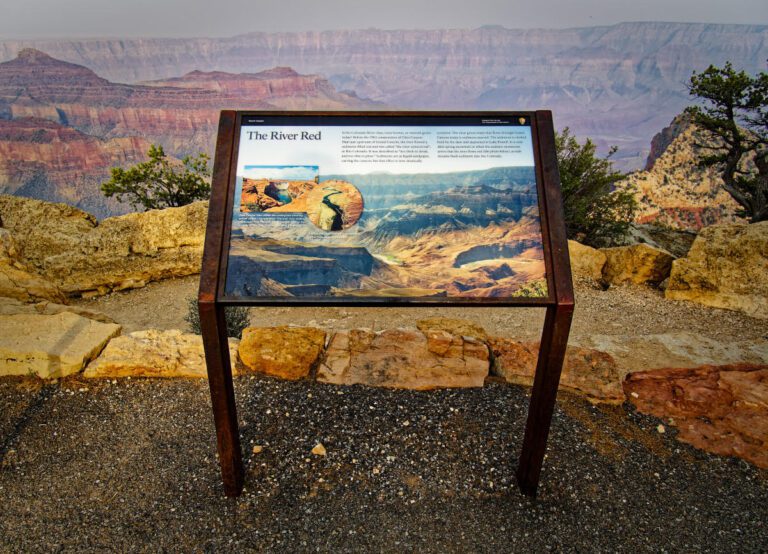A waist high sign with the title "The River Red" depicts the river and rock formations below it. There are two paragraphs on the sign providing information about the river. The sign is perched on an overlook with the river below it and red rock faces in the background.