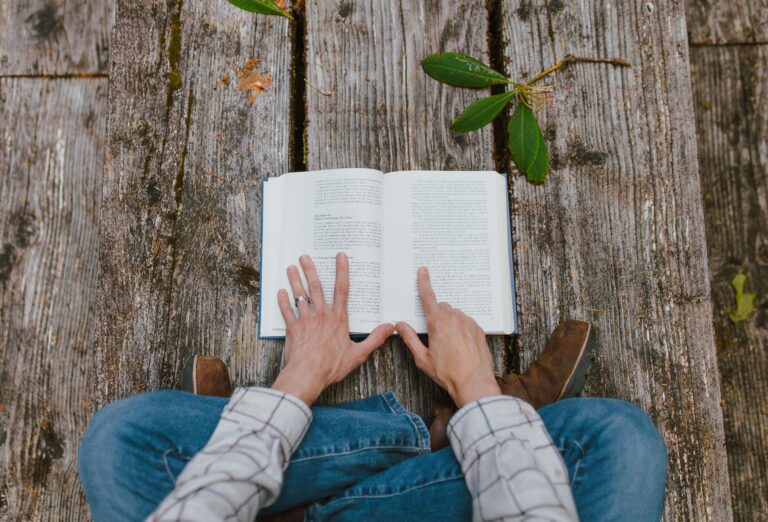 A person sits cross-legged on a wooden deck outside, holding open a textbook with their hands.