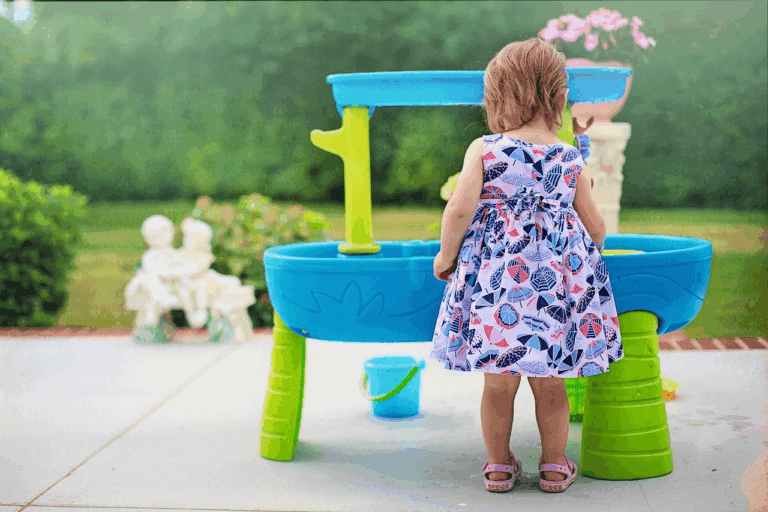 A young child in a dress is standing in the foreground with their back facing the camera. The child is playing with a two story, blue plastic water table that is outside on a cement patio.