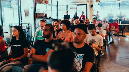 A group of individuals (around 30 are shown) sit in chairs and are focused on a speaker/presentation that is just off screen.