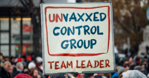 Cardboard sign held during a protest march, saying ‘Unvaxxed control group team leader’.