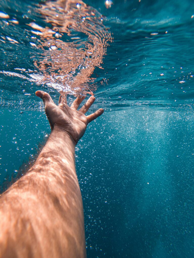 Picture is from the point of view of a person underwater in the ocean. In the foreground, the person's arm is outstretched with their open hand grazing the water surface. Sunlight and bubbles stream around the person;s hand as their fingertips make contact with the surface of the ocean water.
