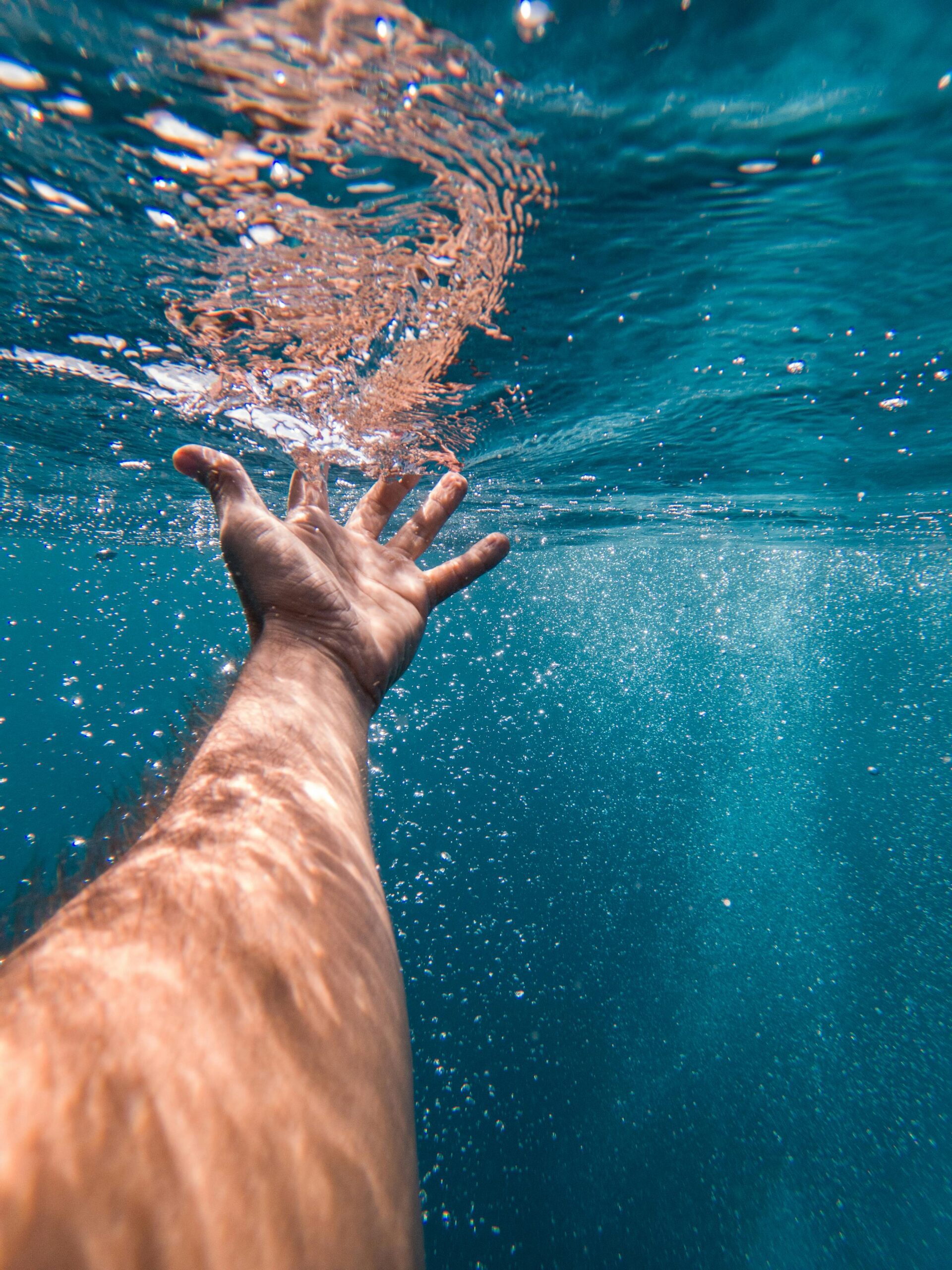 Picture is from the point of view of a person underwater in the ocean. In the foreground, the person's arm is outstretched with their open hand grazing the water surface. Sunlight and bubbles stream around the person;s hand as their fingertips make contact with the surface of the ocean water.