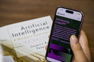Alt text for featured image: Close-up of a Person Holding a Smartphone Displaying ChatGPT. The person is holding the phone in front of a table with a book titled "Artificial Intelligence". The phone screen shows the introduction screen for ChatGPT. The introduction text reads " We’ve trained a model called ChatGPT which interacts in a conversational way. The dialogue format makes it possible for ChatGPT to answer followup questions, admit its mistakes, challenge incorrect premises, and reject inappropriate requests."