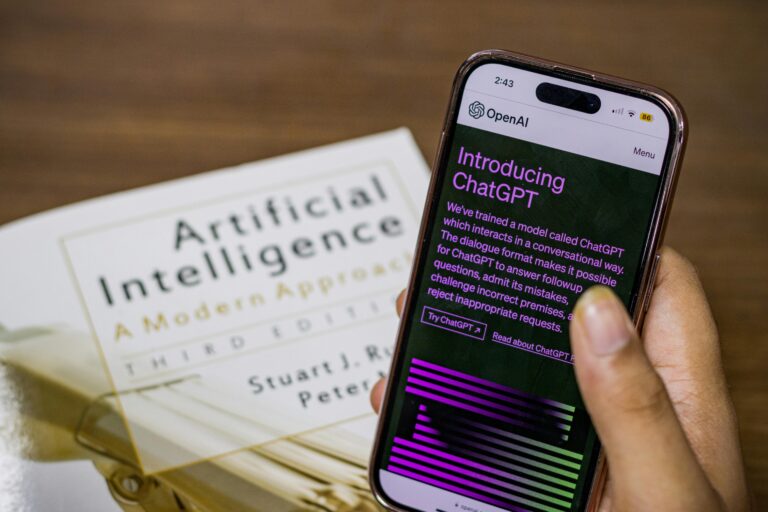 Alt text for featured image: Close-up of a Person Holding a Smartphone Displaying ChatGPT. The person is holding the phone in front of a table with a book titled "Artificial Intelligence". The phone screen shows the introduction screen for ChatGPT. The introduction text reads " We’ve trained a model called ChatGPT which interacts in a conversational way. The dialogue format makes it possible for ChatGPT to answer followup questions, admit its mistakes, challenge incorrect premises, and reject inappropriate requests."