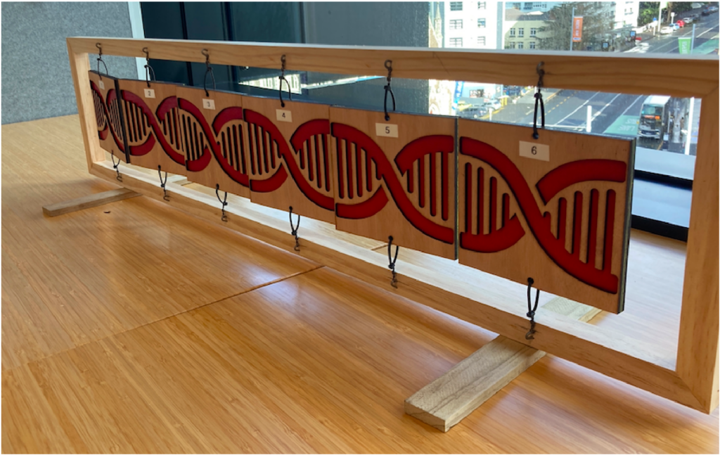 In the centre of the image there is a wooden frame sitting upright on a wooden surface. Suspended within the wooden frame are 6 squares of wood, tied to the top and bottom of the frame so they can spin freely. Across the six squares there is an illustration of the double-helix shape of DNA. There is a window in the background of the image, showing a high-up view of a city road with traffic.