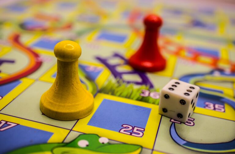 An extreme closeup of the board game snakes and ladders. A yellow player’s pawn and white six sided die are in focus towards the centre of the image. There is a red player’s pawn in soft focus in the background.