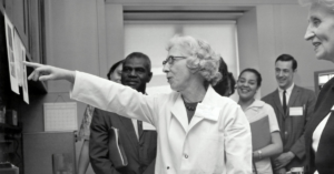 Black-and-white historical photo of a female scientist leading a lab tour, explaining research to a diverse group of attendees.