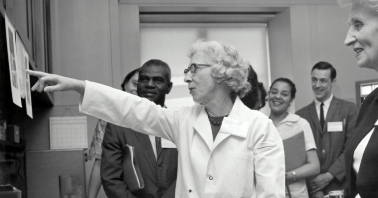 Black-and-white historical photo of a female scientist leading a lab tour, explaining research to a diverse group of attendees.