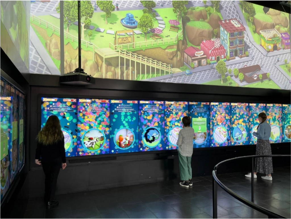 The wall of a dark room is lined with touch screens. There are three people interacting with touch screens spread throughout the room. Each touch screen shows an individual game screen with a blue background and a circle in the centre showing the player character’s environment. Above the touch screens, an isometric map is projected onto the wall, depicting a small town.